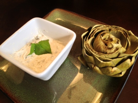 A steamed artichoke and a ramekin with aioli on a plate