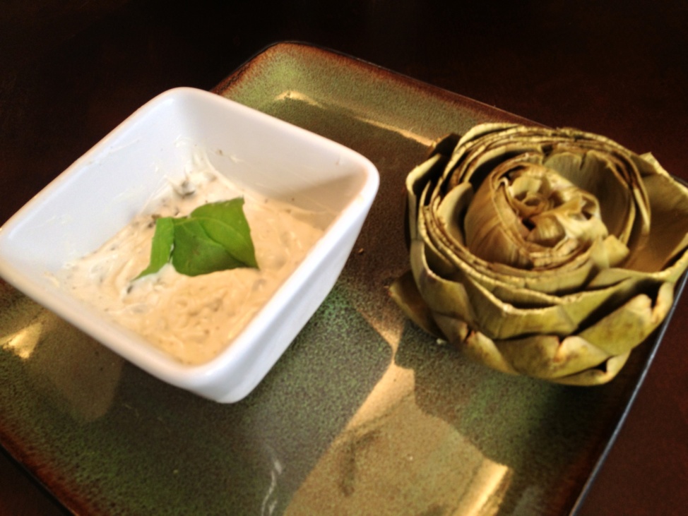 A steamed artichoke and a ramekin with aioli on a plate